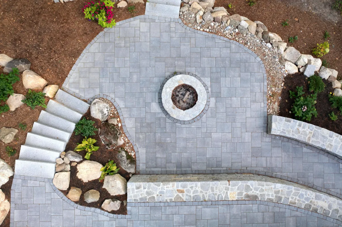 Custom stone patio featuring built-in seating, fire pit, and landscaped borders with plants and stones, designed for outdoor living by Maine Outdoor Spaces.
