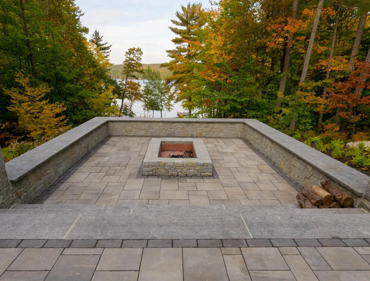Custom stone patio with built-in fire pit, surrounded by autumn foliage and a serene lake view, showcasing outdoor living design by Maine Outdoor Spaces.