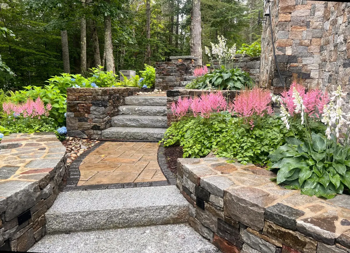 Retaining wall and stone steps surrounded by lush greenery and colorful flowers, enhancing outdoor space design by Maine Outdoor Spaces.